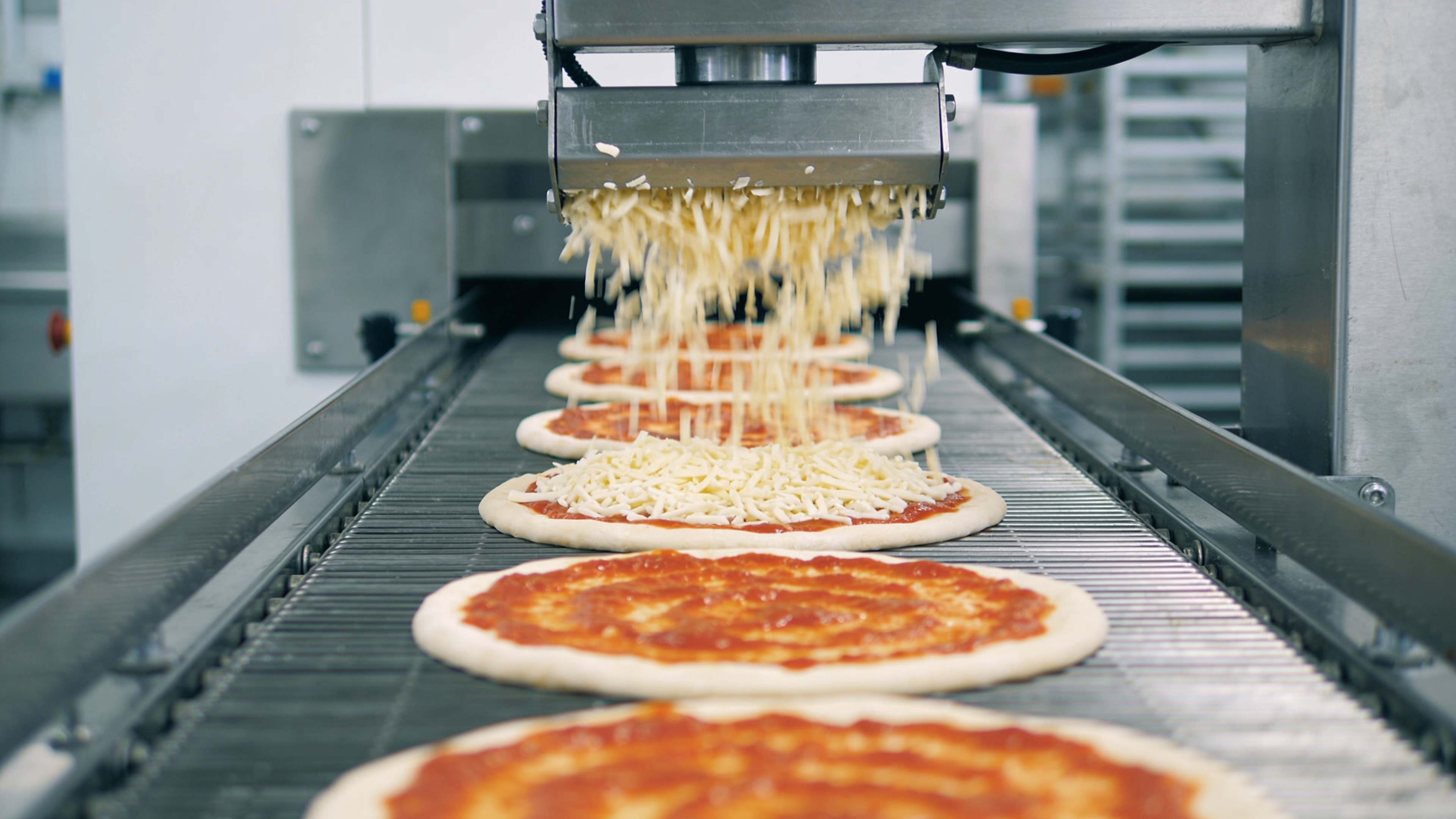 Línea de producción que reparte mozzarella rallada sobre bases de pizza con tomate.